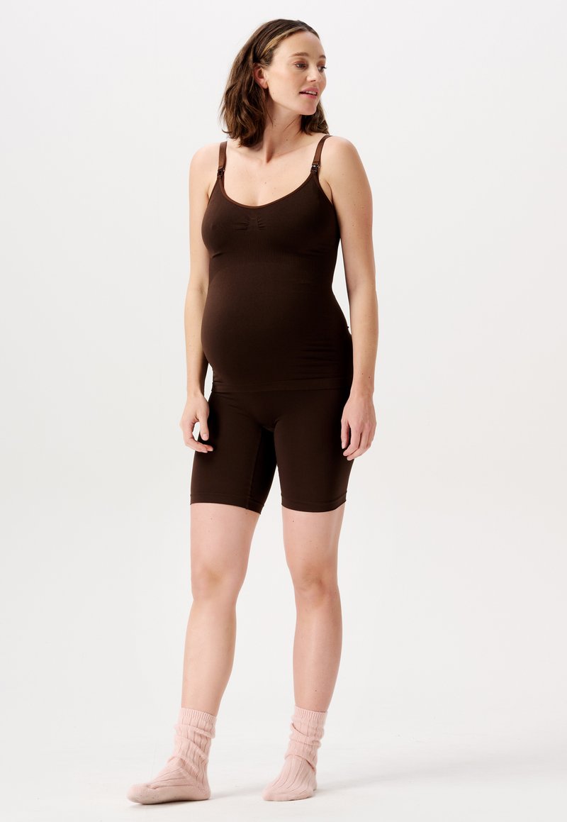 Gravid kvinde iført en mørkebrun ærmeløs top og shorts, stående barfodet med lyserøde sokker på en hvid baggrund.