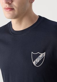 Námořnická modrá tričko s kulatým výstřihem, které má na levé hrudi bílé vyšívané logo ve tvaru štítu, hladký bavlněný textil.