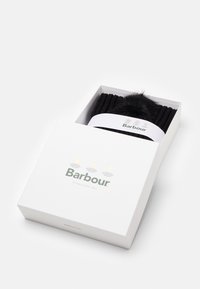 Căciulă tricotată neagră cu un pompon pufoasă într-o cutie albă. Cutia are branding verde și auriu pentru Barbour, înființată în 1894.