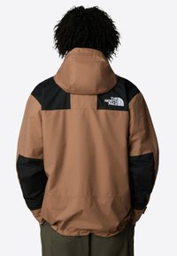 Brązowa wodoodporna kurtka z czarnym wzorem na ramionach i rękawach, z kapturem i logo na plecach. Gładka tekstura z elastycznymi mankietami.