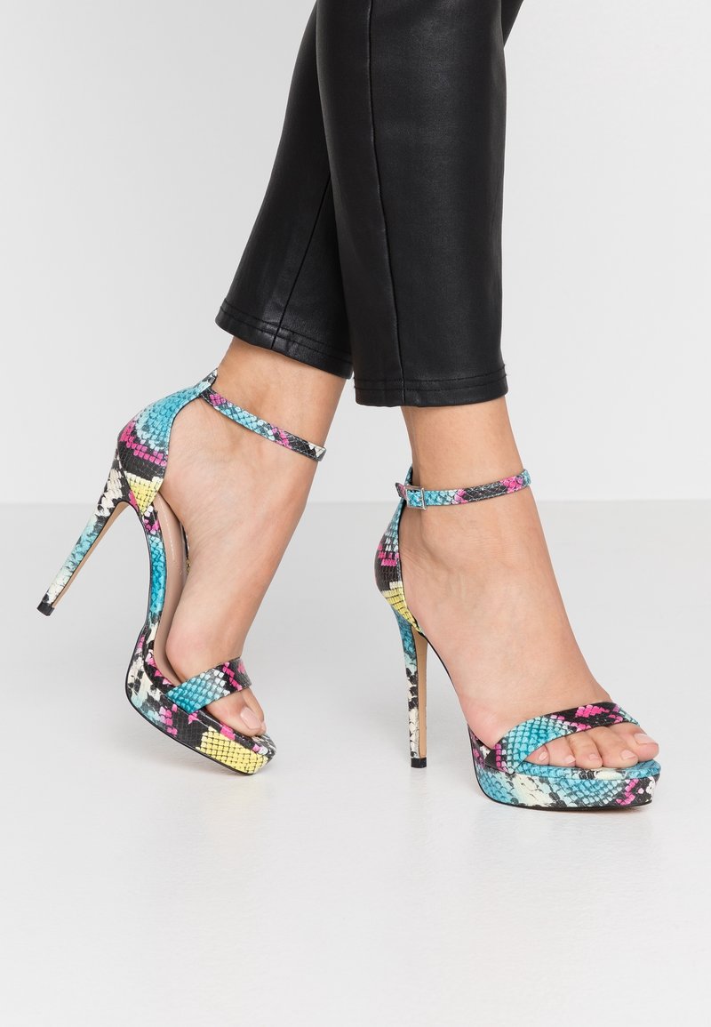 Jambes portant un pantalon noir court et des sandales à talons hauts colorées avec des brides à la cheville et un motif peau de serpent sur un sol blanc.