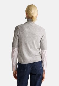 Žena s blond vlasy v nízkém copu má na sobě šedý krátký svetr přes bílou dlouhou košili a tmavě modré džíny, viděná zezadu.