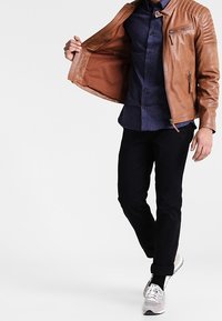 Giacca da biker in pelle marrone con spalle rigate, indossata aperta su una camicia a fantasia blu navy, abbinata a pantaloni neri e sneakers grigie chiare.