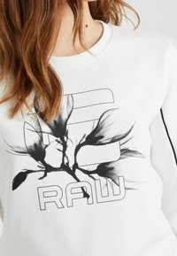 Sudadera blanca con un gráfico floral negro y el texto "RAW" en negrita en el centro. Textura suave y cuello redondo.