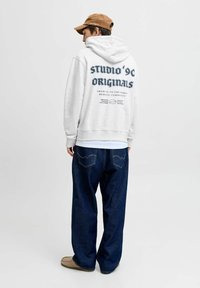 Sudadera gris con un gran estampado gráfico en la parte trasera, combinada con jeans de corte suelto en azul oscuro y calzado claro, sobre un fondo simple.
