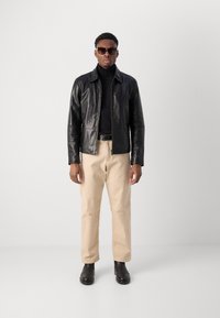 Μαύρο δερμάτινο μπουφάν με κλείσιμο με φερμουάρ, φορεμένο πάν ω από ένα μαύρο ζιβάγκο, σετ με μπεζ παντελόνι και μαύρα μποτάκια αστραγάλου. Αξεσουάρ περιλαμβάνουν γυαλιά ηλίου.