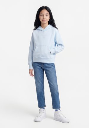 Κορίτσι με μακριά μαύρα μαλλιά, που φοράει ανοιχτό μπλε φούτερ με κουκούλα, μπλε τζιν και λευκά αθλητικά παπούτσια, στέκεται με το ένα χέρι στην τσέπη.