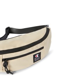 Riñonera beige hecha de un tejido duradero con una textura suave. Presenta un cierre negro, una correa ajustable y un parche de logo en el lateral.