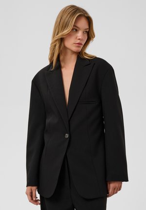 Mujer con cabello castaño claro que lleva un blazer negro de un solo botón y talla grande, de pie frente a un fondo claro y liso.