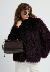 Kvinde med krøllet hår, iført gyldne hoop øreringe, mørk fluffy jakke, solbriller, cremede bukser og holder en mørkebrun læder taske.