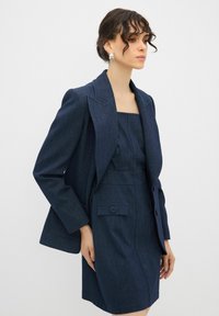 Dunkelblaues Jeansblazer und eng anliegendes Kleid aus passendem Stoff, mit quadratischem Ausschnitt, strukturierten Schultern und Knopfdetails am Kleid.