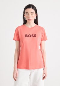 Sieviete ar garām tumšām matiem valkā korallkrāsas T-kreklu ar uzrakstu "BOSS" uz pašas priekšpuses, kuru papildina lielas, apaļas auskari.