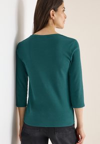 Frau mit langen dunklen Haaren, die eine türkisfarbene 3/4-Ärmel-Bluse und dunkle Jeans trägt, steht und blickt an eine hellfarbige Wand.