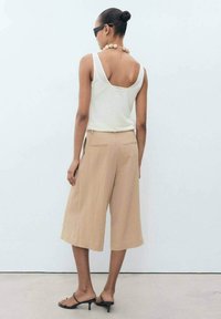 Beige culottes dengan potongan lebar, dipadukan dengan atasan tank berwarna krim yang memiliki punggung melengkung. Aksesorisnya mencakup kalung manik-manik bulat dan kacamata hitam.