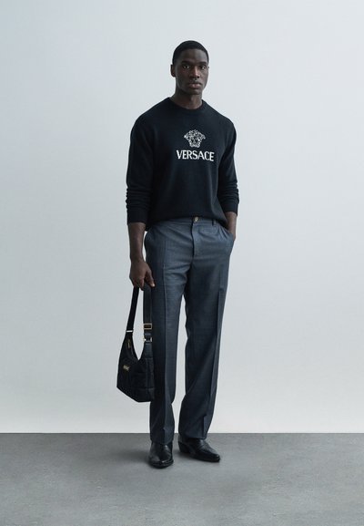 Černý pletený svetr s logem "VERSACE", šedé upravené kalhoty a černá prošívaná kabelka. Nošeno s černými koženými botami s minimálním designem.