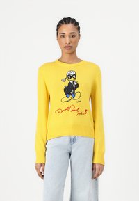 Pull en maille jaune avec un graphique d'un canard de dessin animé portant des lunettes, avec un texte rouge en dessous. Manches longues et col rond.