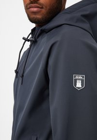 Giacca a vento blu navy con una texture liscia, dotata di un cordino regolabile e di un logo prominente sul manica sinistra.