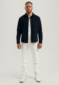 Marineblauw textuurshirt met knoopsluiting en zak, gecombineerd met witte rechte jeans en witte sneakers. Model staat tegen een neutrale achtergrond.