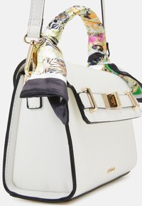 Valkoinen rakenteellinen käsilaukku, jossa mustat reunat, teksturoitu pinta, kullanväriset metalliosat ja monivärinen kuvioitu huivi sidottuna kahvaan.