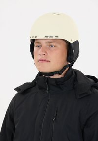 Hvid skihjelm med ventilationsåbninger, båret sammen med en sort jakke med lynlås og velcrolukninger. Enkel design, mat finish.