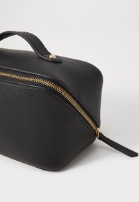 Borsa cosmetica in pelle nera texture con zipper dorato, forma rotonda, manico superiore e un piccolo tirante sulla zip.