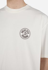 Tricou alb din bumbac cu guler rotund, având un logo circular de culoare maro închis care spune "DOLCE VITA PEGADOR." Mâneci scurte.