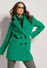 Groene dubbelzijdige jas met een maatwerkontwerp, grote zwarte knopen en zijzakken. Gestyled met een groene coltrui en zonnebril.