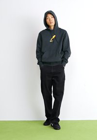 Mørkegrå hættejakke med en frontlomme og et lille gult grafisk design. Sorte baggy bukser og sorte sneakers, stående mod en hvid baggrund.