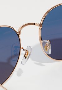 Ochelari de soare din metal cu nuanță de auriu, cu lentile de un albastru închis, design în formă de inimă și pernițe de nas clare. Prezintă un detaliu mic cu logo pe pernă.