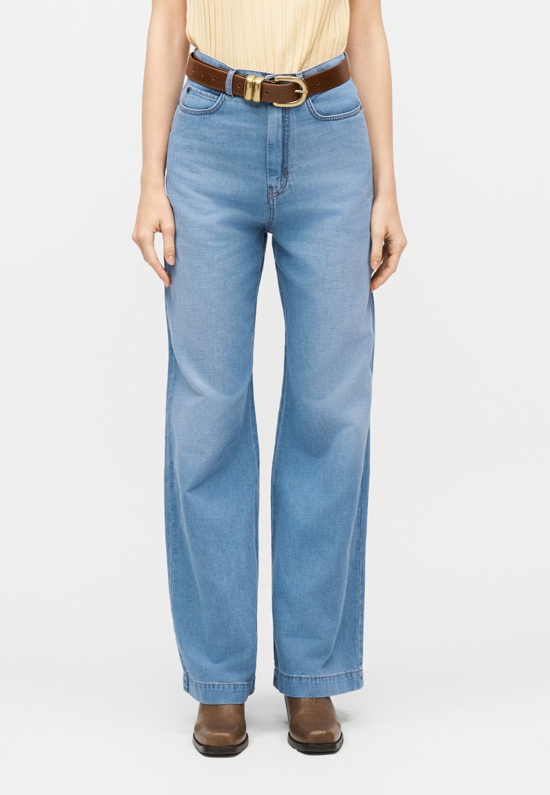 Femme portant un jean large bleu clair, une ceinture marron avec une boucle dorée et des chaussures marron, debout devant un fond uni.