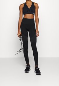 Sujeción deportiva negra con un diseño cruzado, combinada con leggings negros de tiro alto que presentan paneles texturizados. Sostiene una cuerda de saltar con una mano.