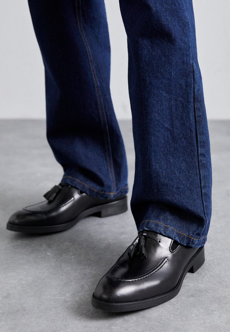 Mocassins en cuir noir avec un détail de frange, associés à un jean en denim bleu. Les chaussures ont un bout rond et un petit talon carré.