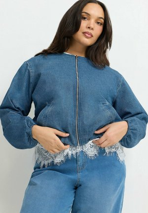 Femme portant une veste en jean bleue zippée avec des manches bouffantes sur un top en dentelle blanche et un jean en denim bleu assorti.