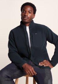 Dark green fleece jacket with a high collar, zip closure, and a chest pocket. Model seated on a stool against a neutral background.