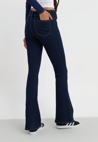 Mørkeblå utsvingte jeans med en jevn tekstur, som har to baklommer og kontrasterende sømmer, sammen med svart og hvite sneakers.