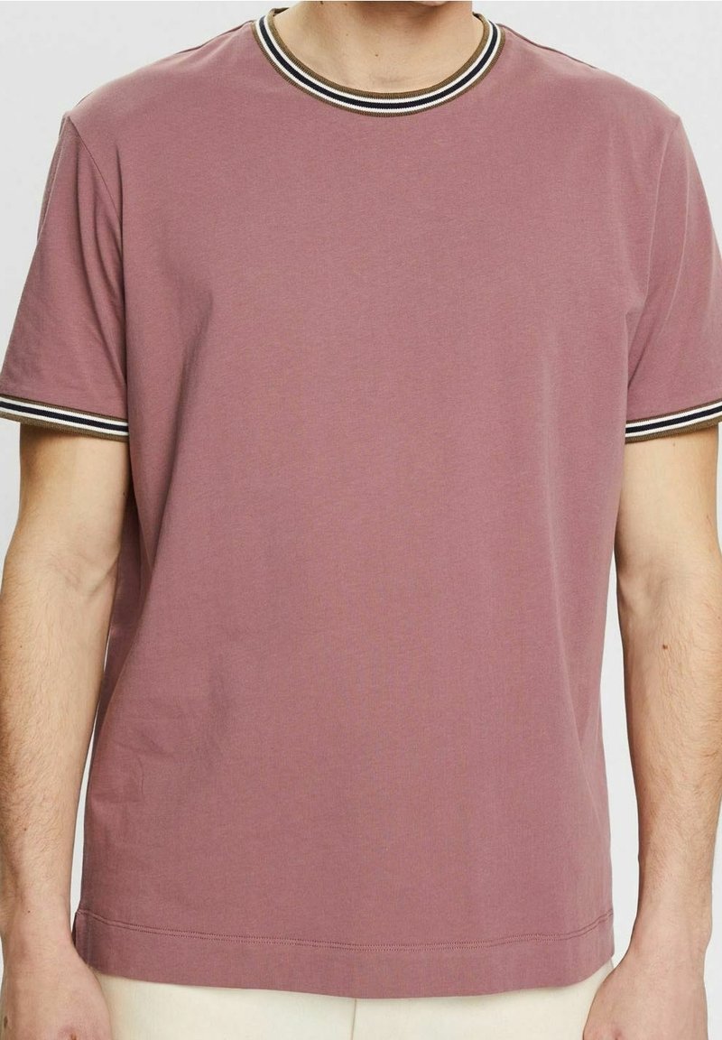 Homme portant un t-shirt mauve à manches courtes avec un col et des poignets côtelés rayés noir, blanc et beige, debout devant un fond uni.
