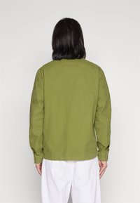 Khaki grön långärmad jacka, tillverkad av bomull. Har en avslappnad passform, rak fåll, knappmanschetter och minimalistiska sömdetaljer.