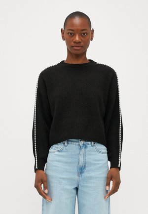 Pull en maille côtelée noir avec un col ras du cou, des épaules tombantes et des rayures latérales avec du détail blanc. Porté avec un jean bleu clair.