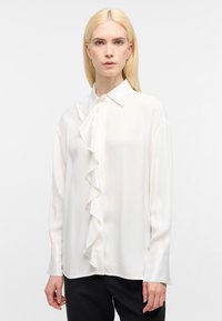 Blusa bianca a maniche lunghe con dettagli arricciati nella parte anteriore, tessuto morbido e colletto. Texture liscia, abbinata a pantaloni neri.