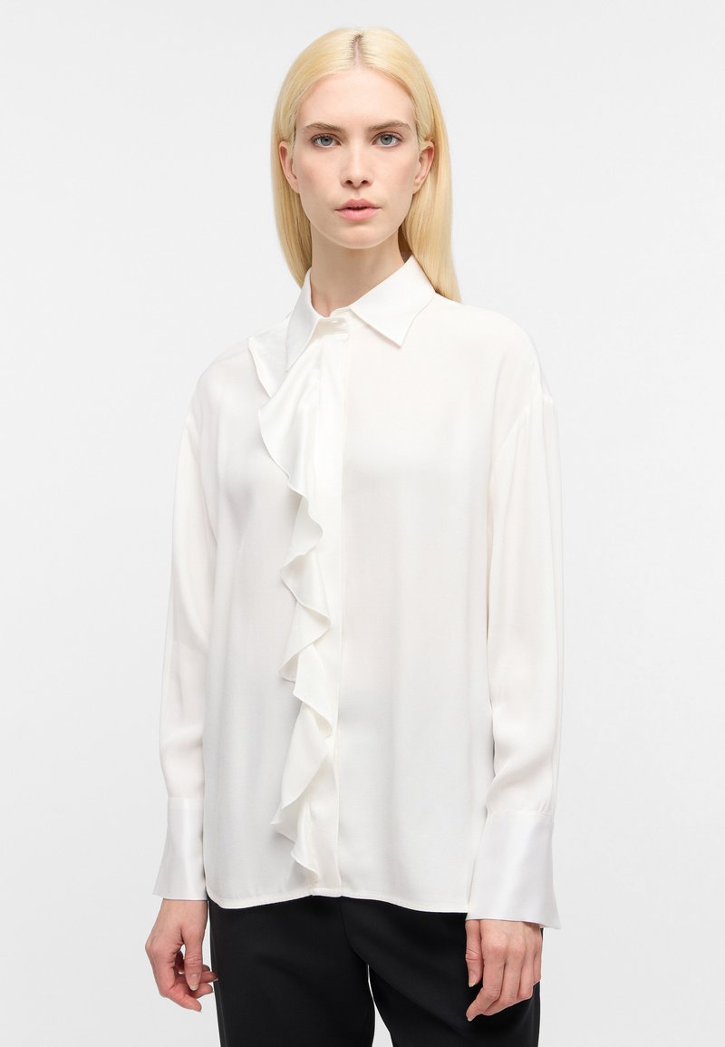 Blusa bianca a maniche lunghe con dettagli arricciati nella parte anteriore, tessuto morbido e colletto. Texture liscia, abbinata a pantaloni neri.
