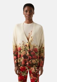 Kardigan w kremowym kolorze z kwiatowym wzorem, o prążkowanej fakturze, z otwartym przodem i dużymi guzikami, zestawiony z pasującymi spodniami w nadruk.