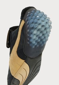 Black and tan sports shoe with textured leather, reinforced toe cap, and a patterned rubber sole featuring blue cushioning and branding.