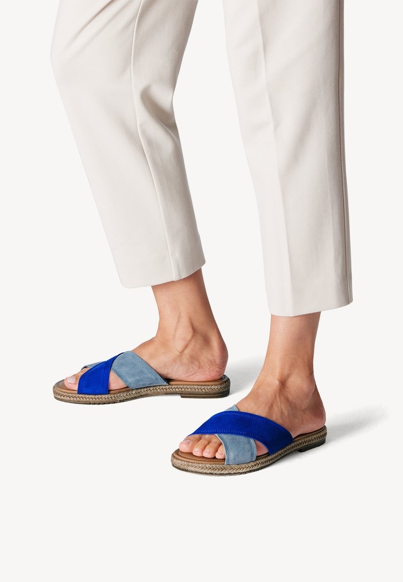 Fötter i slip-on sandaler med blå och ljusblå mockakorsade remmar och sula med repdetaljer, ihop med krämfärgade avklippta byxor.