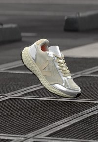 Sportowe sneakersy w kremowej i srebrnej siateczce z teksturowaną gumową podeszwą; odblaskowe elementy i minimalistyczny design. Zawierają sznurówki i akcenty po bokach.
