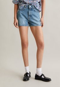 Lichte blauwe denim shorts met een hoge taille, klassiek vijf-pocket ontwerp en een rafelzoom, gecombineerd met zwarte schoenen en witte sokken.