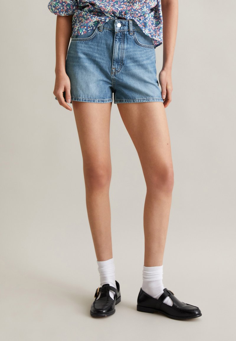 Lichte blauwe denim shorts met een hoge taille, klassiek vijf-pocket ontwerp en een rafelzoom, gecombineerd met zwarte schoenen en witte sokken.