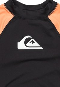 Rash guard negro y naranja, textura suave, cuello alto, costuras planas, presenta un logo blanco con un diseño de montaña y ola.