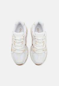 Białe sneakersy z beżowymi akcentami, z teksturowaną cholewką, sznurowanym zapięciem, wyściółką na kołnierzu i masywną gumową podeszwą. Widoczny detal z logo w kolorze złotym.