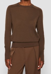 Brązowy sweter dziergany z okrągłym dekoltem, ściągaczami na mankietach i u dołu. Charakteryzuje się subtelną fakturą i dopasowanym krojem, w zestawie z dopasowanymi spodniami.