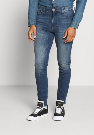 Vaqueros de mezclilla oscuros ajustados con un ligero desgaste, diseño de cinco bolsillos y cierre de cremallera, combinados con zapatillas negras y blancas.